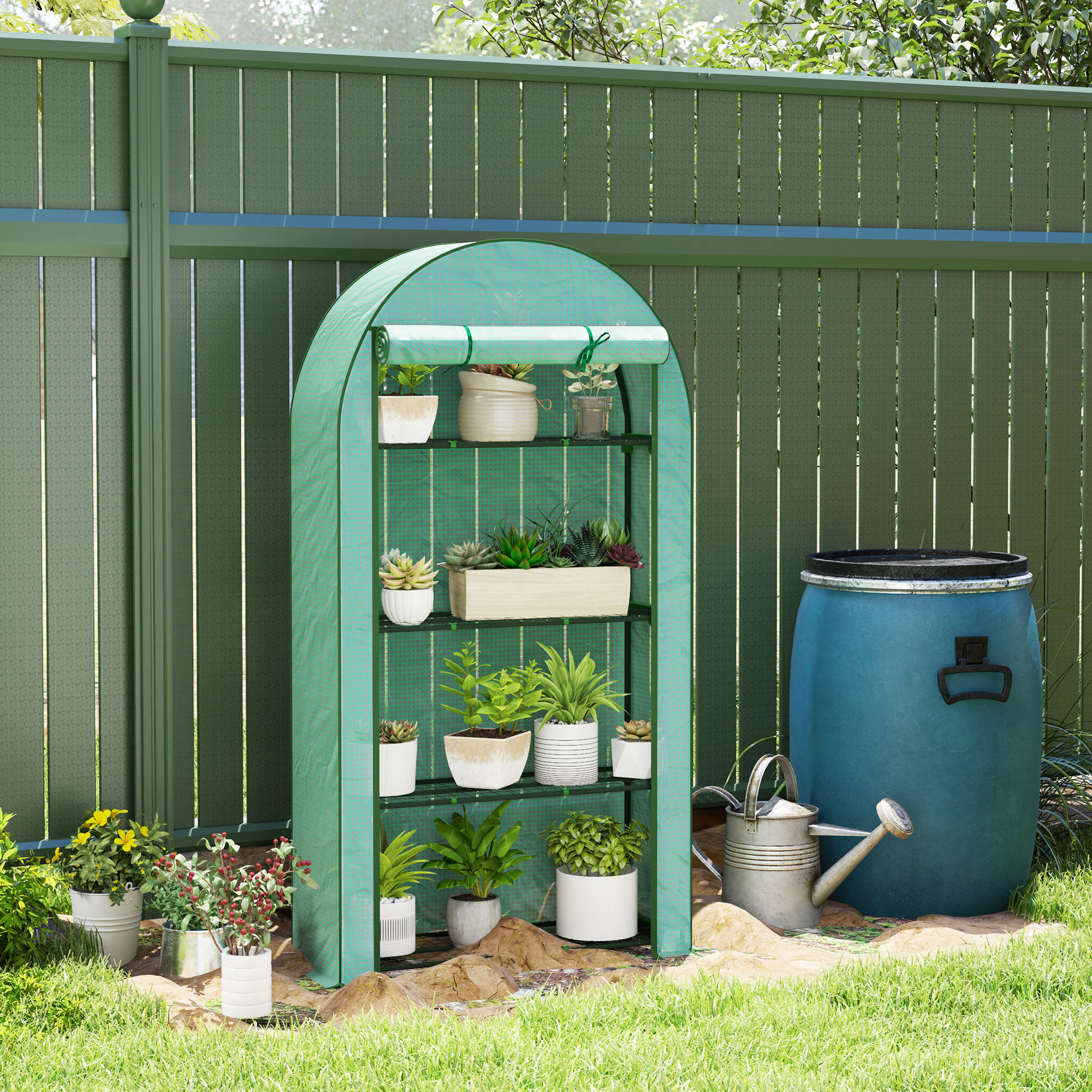 Un mini invernadero verde con plantas se sienta junto a un barril azul y regadera.