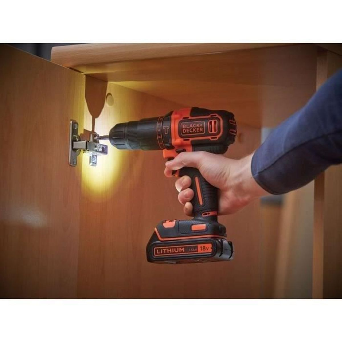 Eine Hand benutzt einen schwarzen und orangen Bohrer, um eine Scharnierschraube in Holz zu drehen.