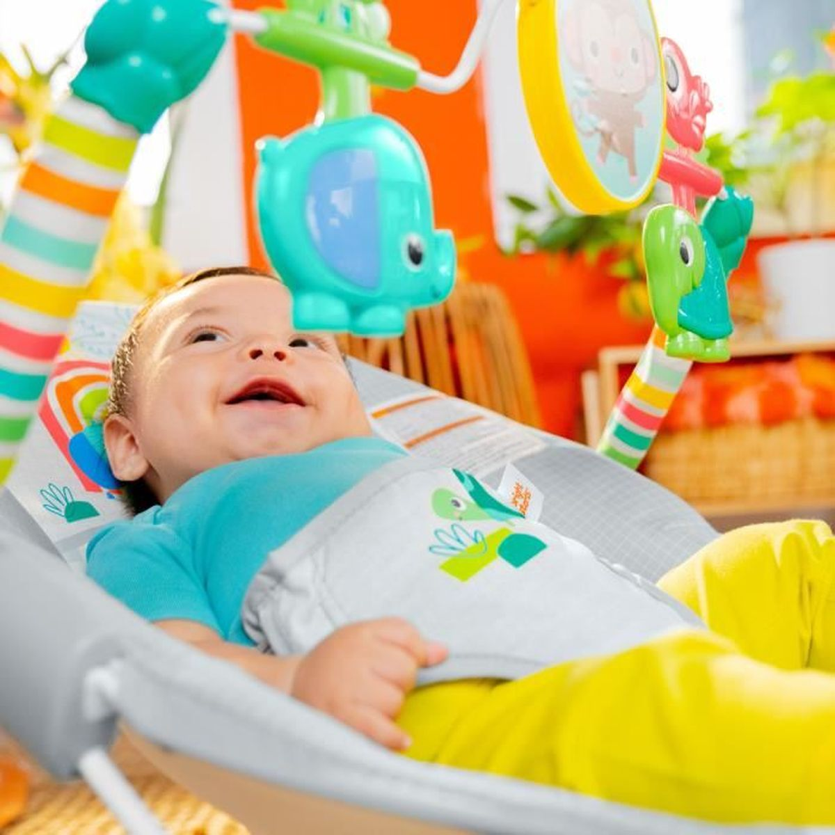 Ein Baby liegt in einer Wippe unter bunten Spielzeugen, darunter ein Elefant.