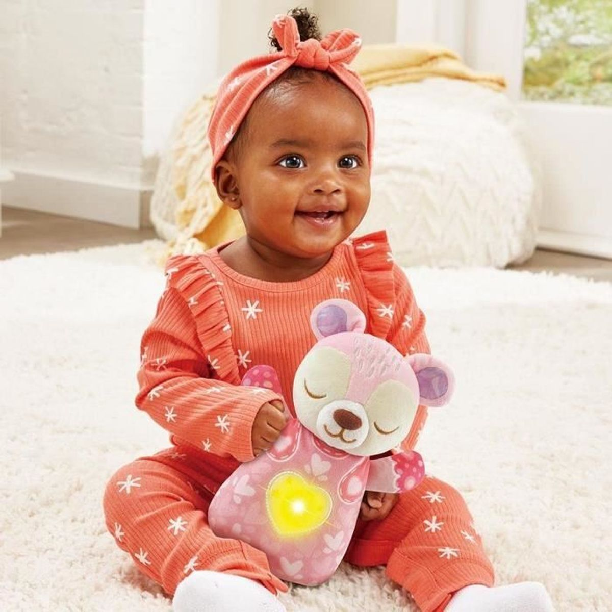 Ein Baby in einem Korallen-Outfit hält ein rosa Teddybär-Spielzeug mit leuchtendem Herzen.