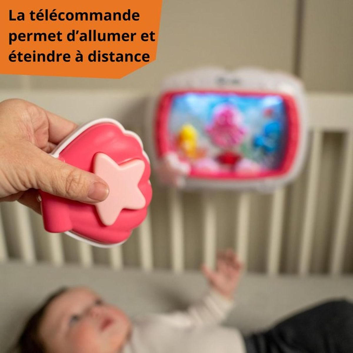 Eine Hand hält eine rosa Fernbedienung mit Stern. Ein Baby liegt im Kinderbett.