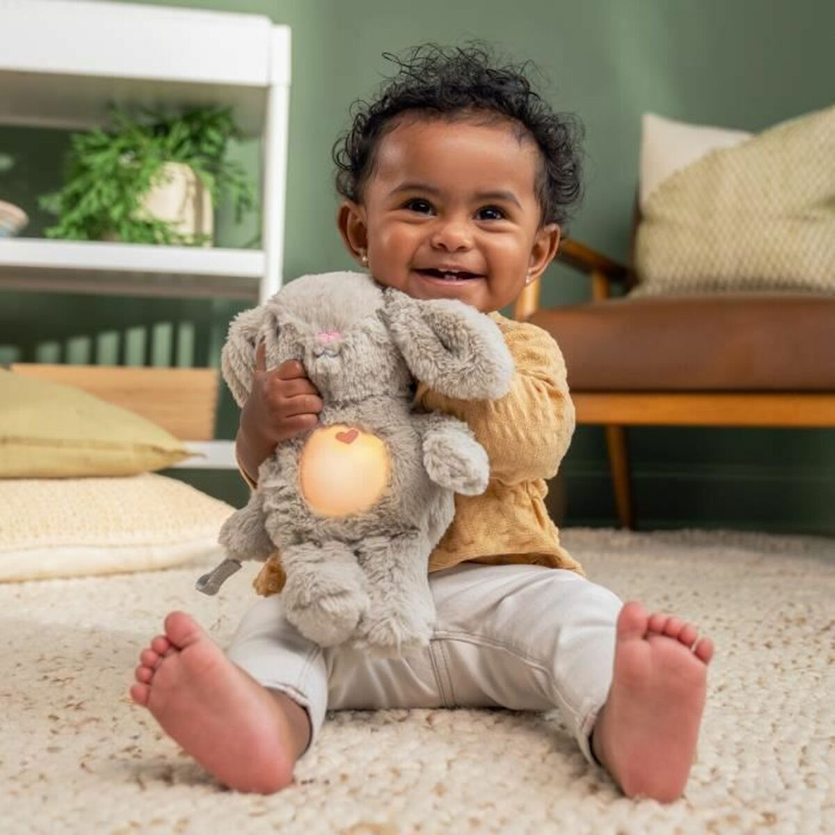 Lächelndes Baby im gelben Hemd hält ein leuchtendes Hasenspielzeug. Barfuß.