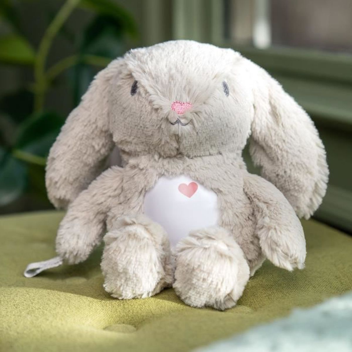 Ein graues Häschen-Stofftier mit rosa Herz auf Bauch und Nase sitzt auf einem grünen Kissen.