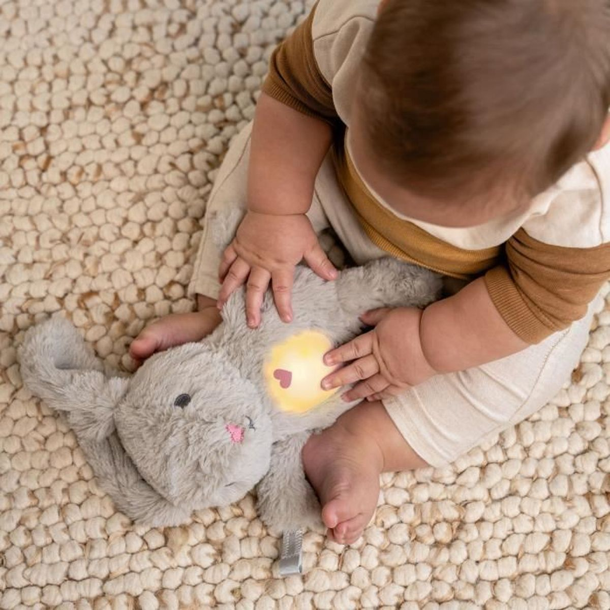 Ein Baby spielt mit einem grauen Hasen-Kuscheltier mit leuchtendem Herz auf einem Teppich.