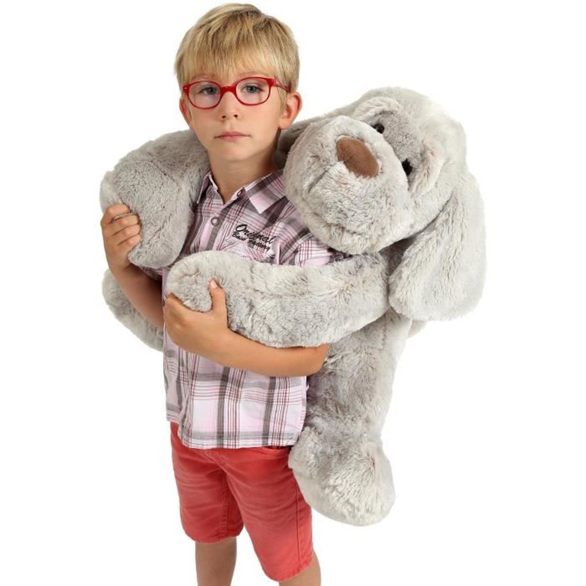 Un garçon avec des lunettes rouges tient un grand chien en peluche gris.