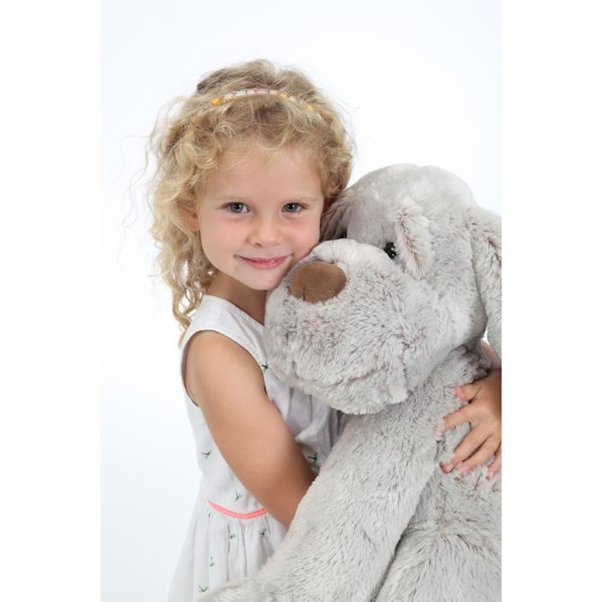 Une petite fille aux cheveux blonds bouclés serre un grand chien en peluche gris.