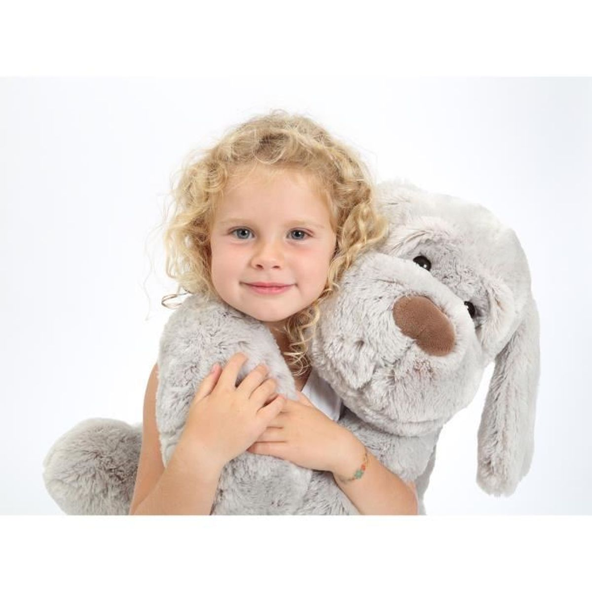 Une petite fille aux cheveux blonds bouclés serre un grand chien en peluche gris.
