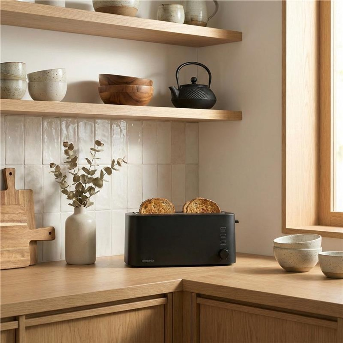 A black toaster with two slices of toast sits on a wooden counter next to a vase with branches.