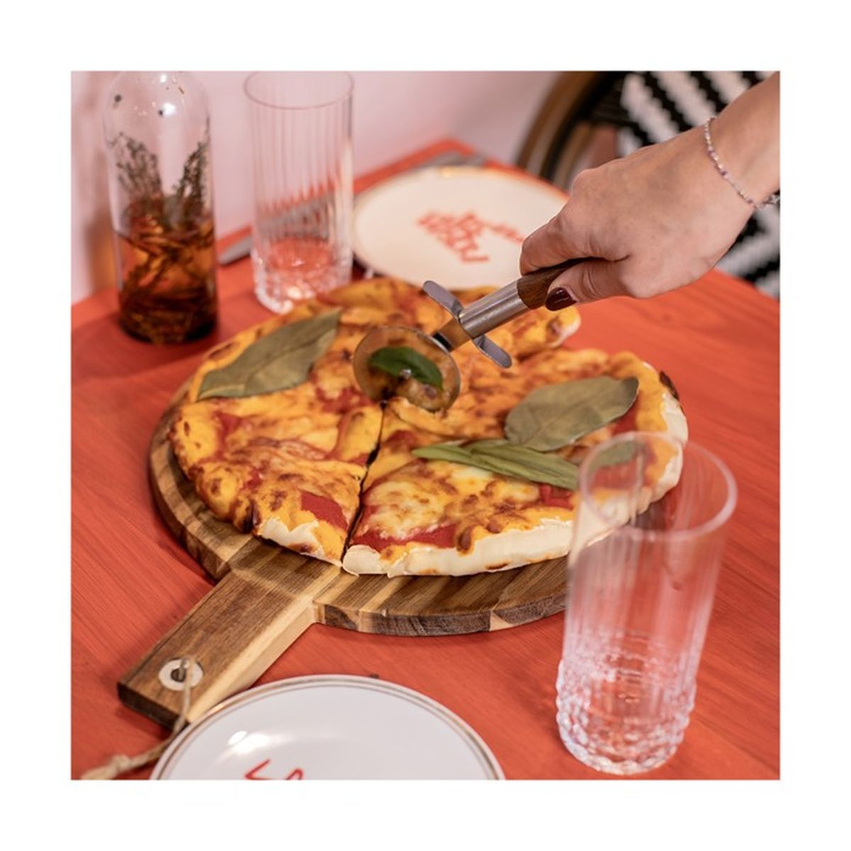 Eine Hand schneidet eine Pizza mit einem Pizzaschneider. Auf dem Tisch stehen Gläser und eine Flasche.