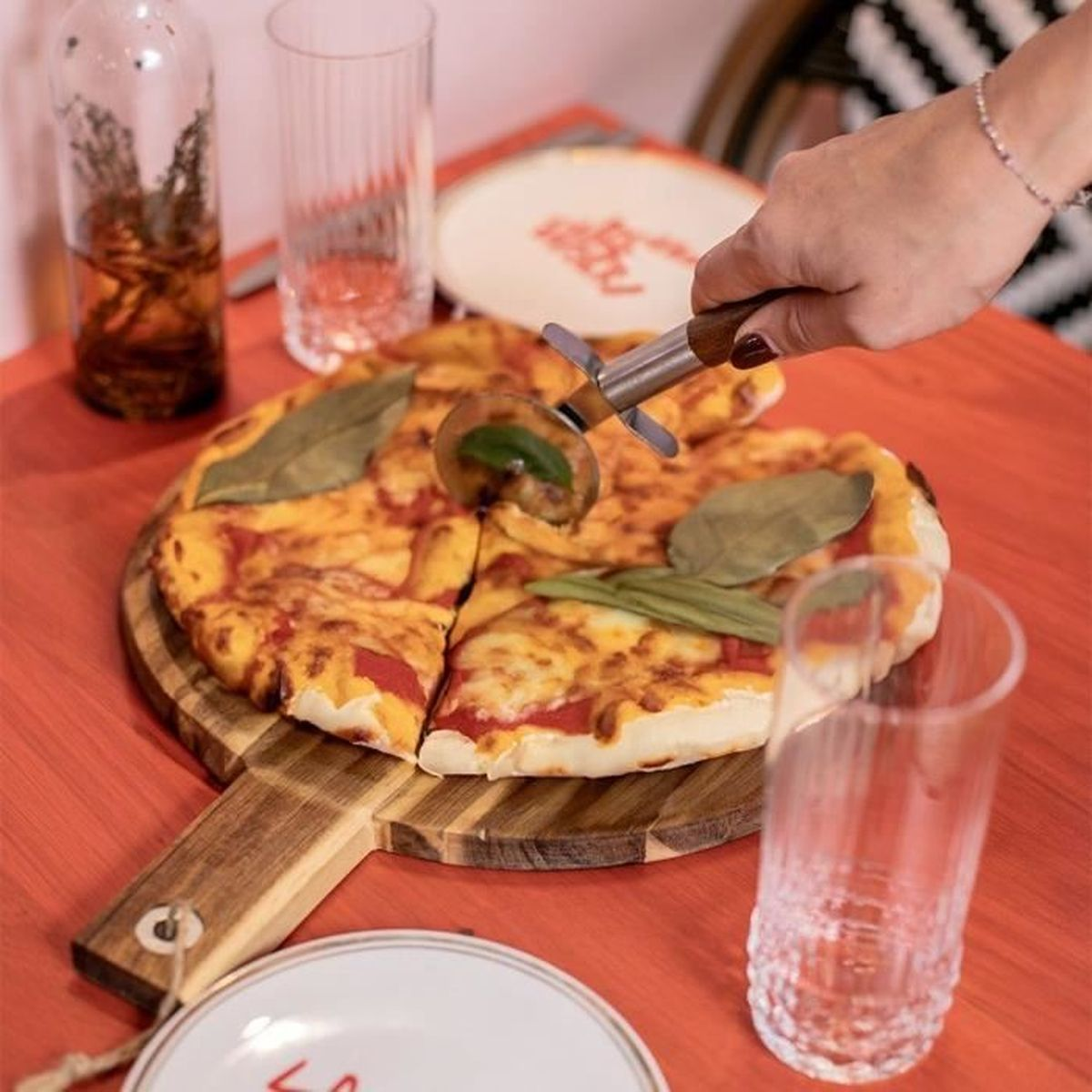 Eine Hand schneidet eine Pizza mit einem Pizzaschneider auf einem Holzteller.