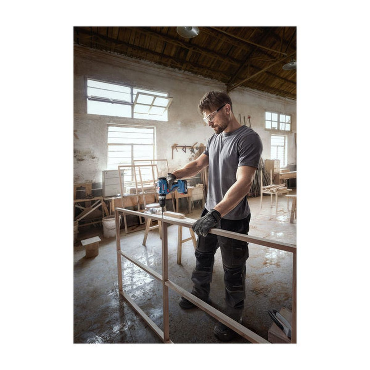 Mann bohrt Holzrahmen in Werkstatt. Er trägt Schutzbrille und Handschuhe.
