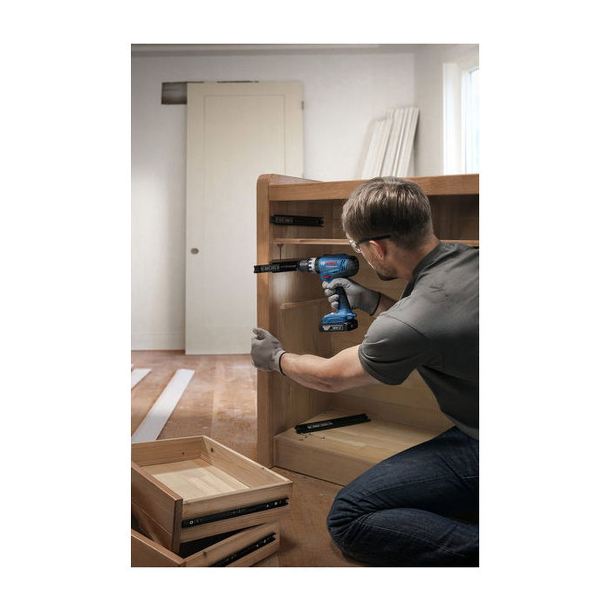 Mann mit Bohrmaschine montiert Holzschrank, Schubladen liegen daneben.