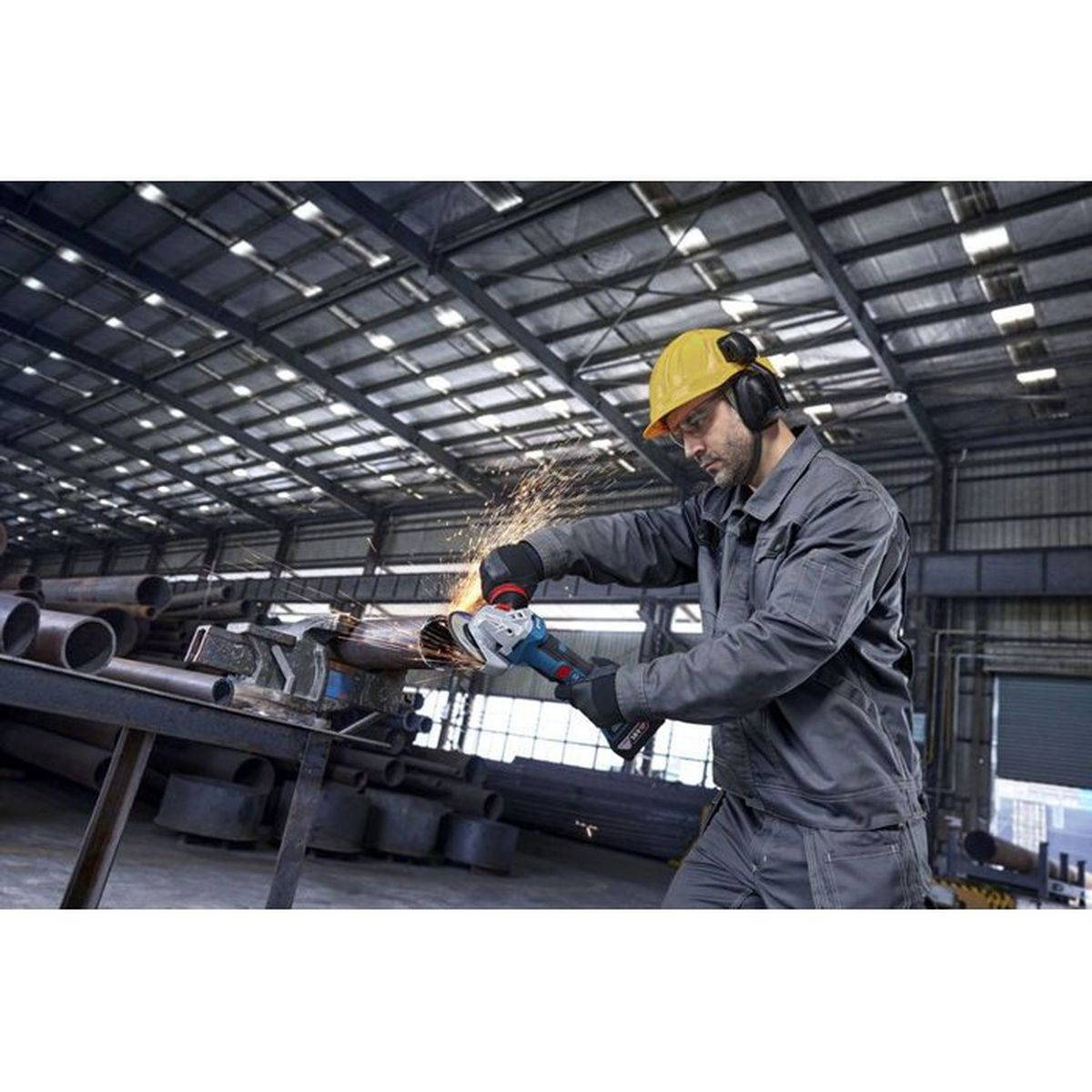Mann mit Helm und Gehörschutz schleift Metallrohr; Funken sprühen.