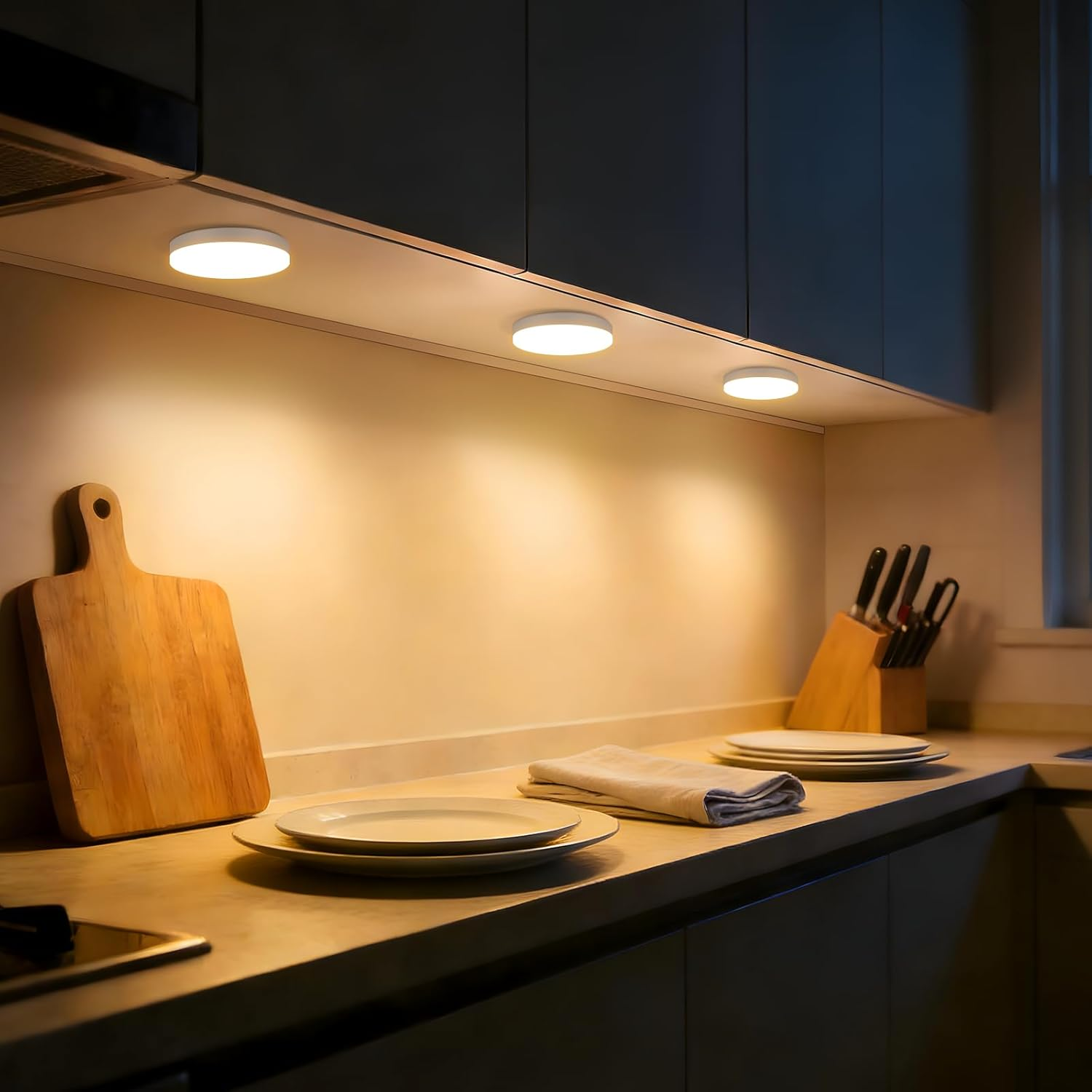 Una cucina con piano di lavoro, tagliere in legno, piatti, ceppo coltelli e luci sottopensile.