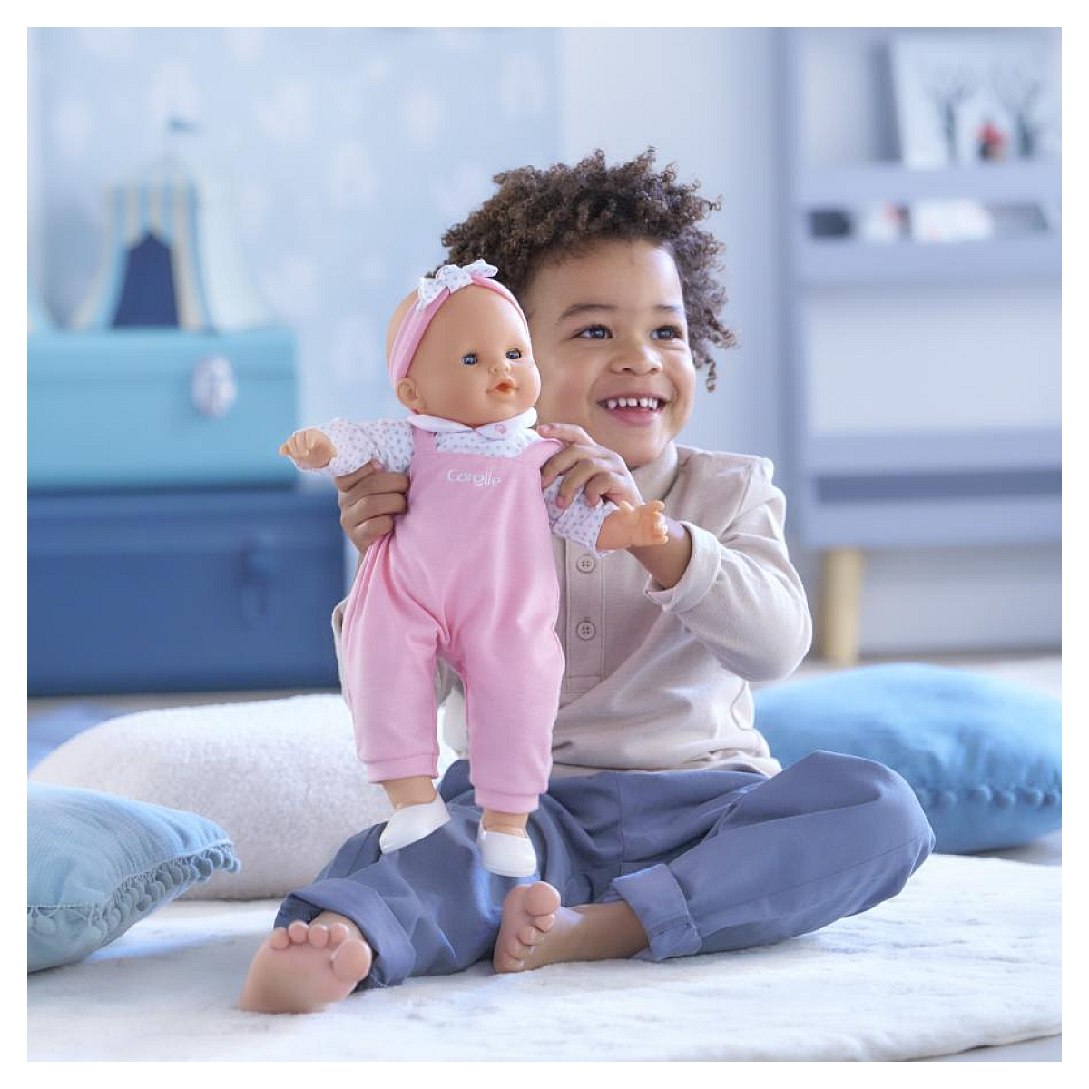Ein Junge hält eine Puppe in rosa Latzhose und spielt auf einem Teppich.