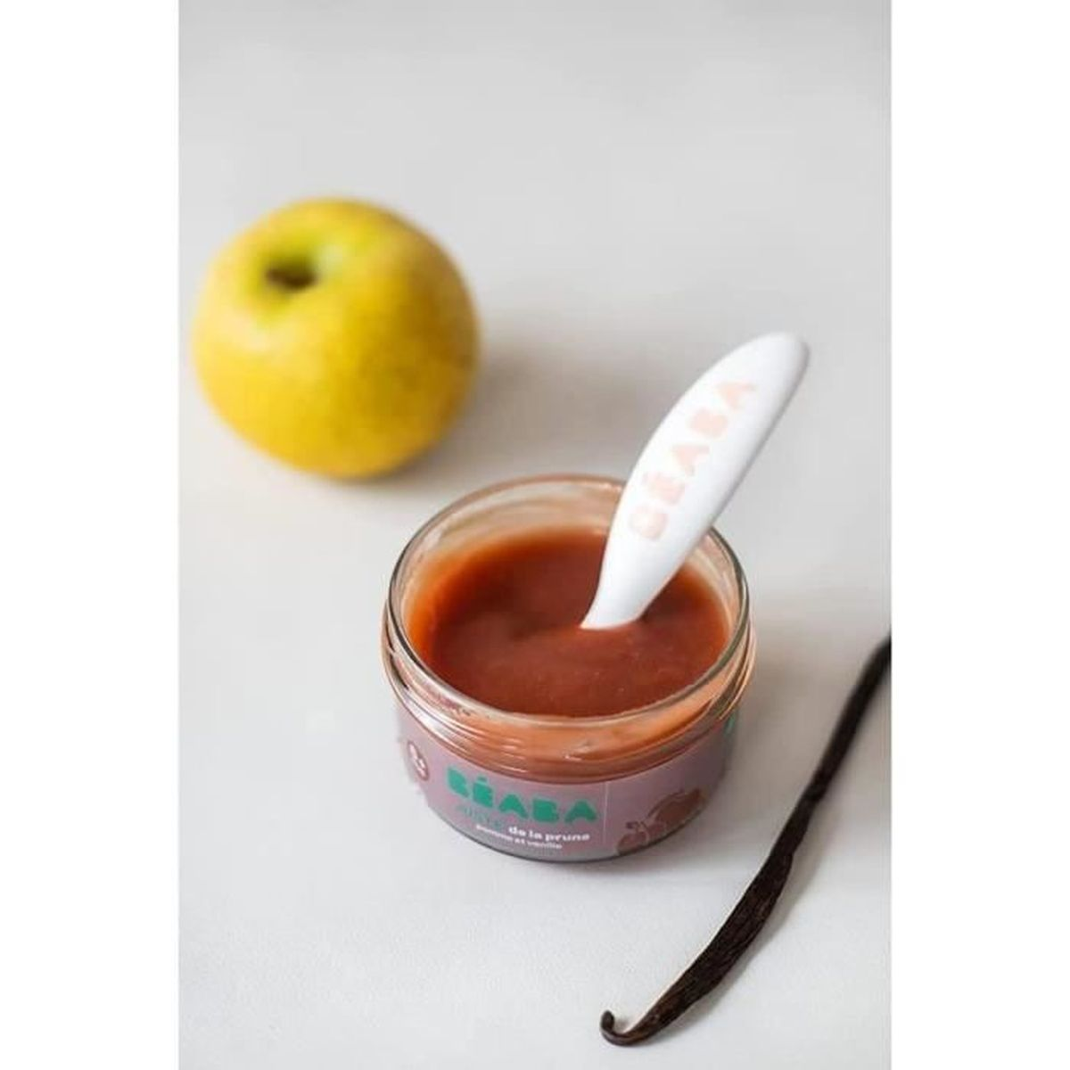 Ein Glas Pflaumenpüree mit Löffel, daneben ein Apfel und eine Vanilleschote.