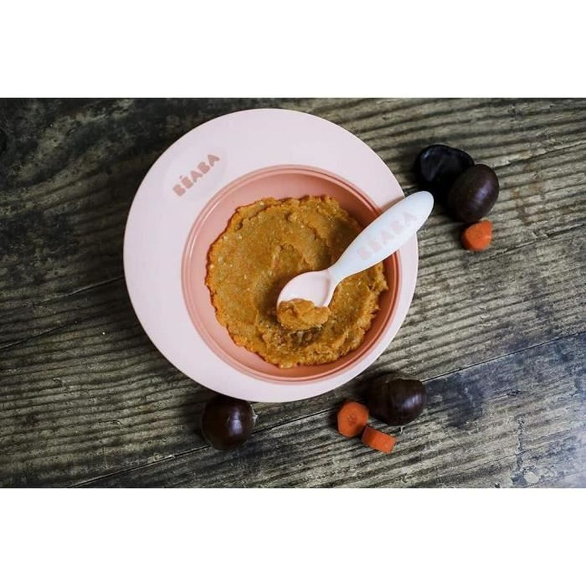 Ein Beaba-Teller mit orangefarbener Babynahrung und einem Löffel. Karottenstücke und dunkle Früchte daneben.