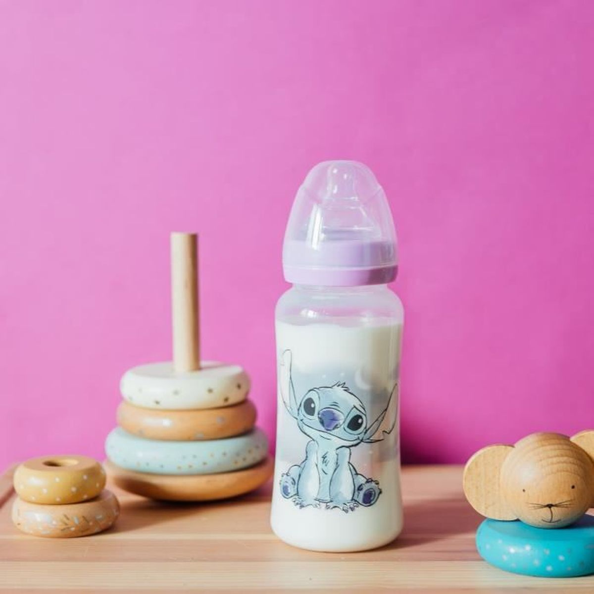 Milchflasche mit Stitch-Motiv neben Holzspielzeug vor pinkem Hintergrund.