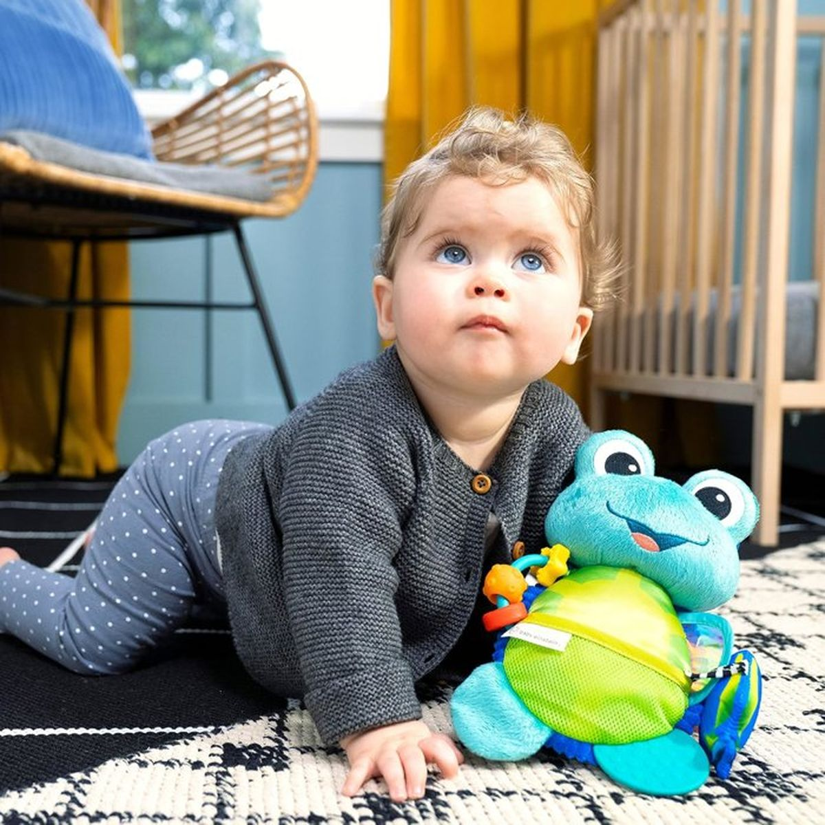 Ein Baby mit blauen Augen krabbelt auf einem Teppich neben einem Froschspielzeug. Ein Bettchen im Hintergrund.