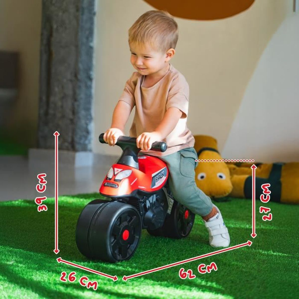Un bambino cavalca una moto giocattolo rossa su erba sintetica. Vengono mostrate le dimensioni.