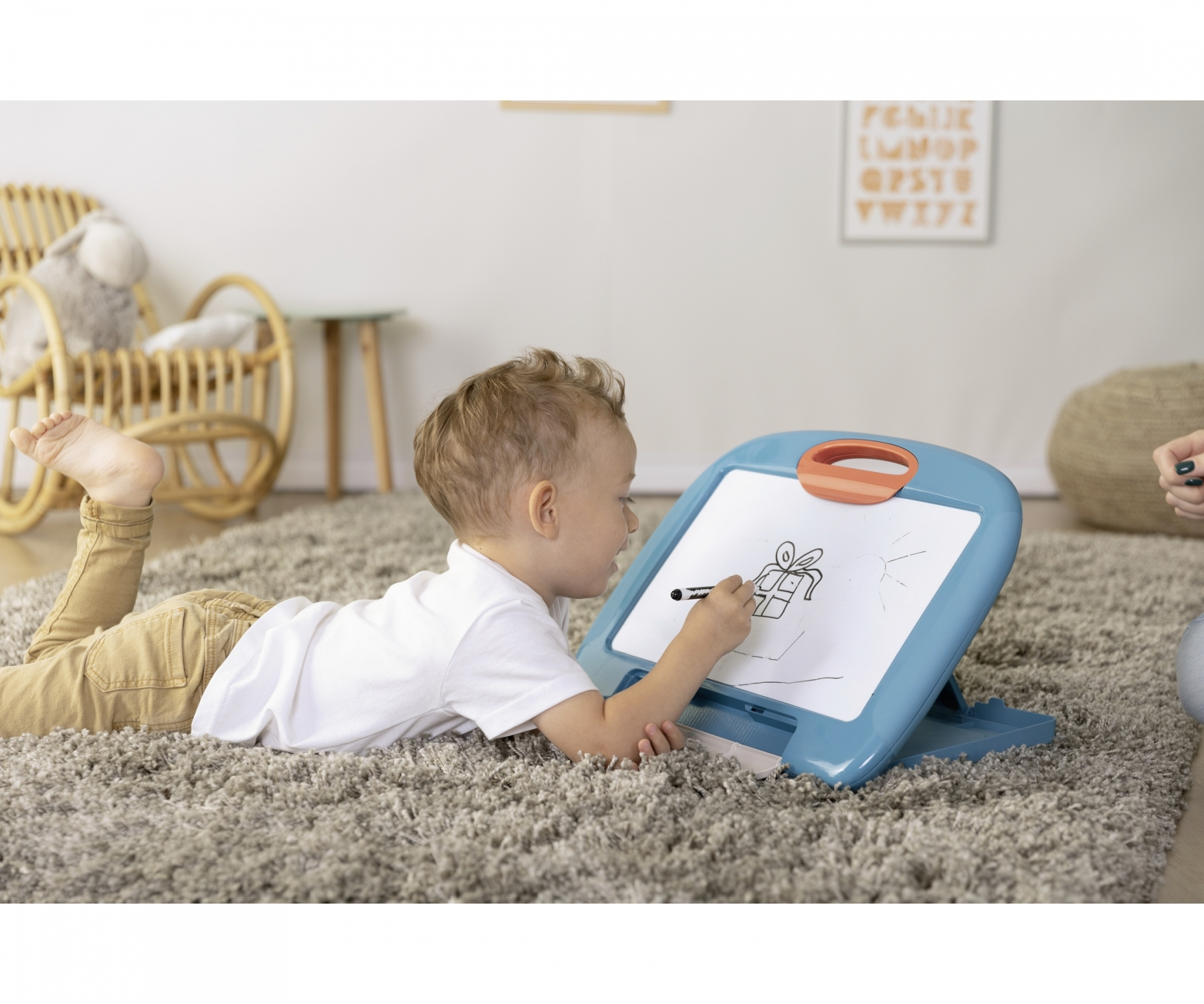 Un bambino piccolo giace su un tappeto, disegnando un regalo su una lavagna blu.