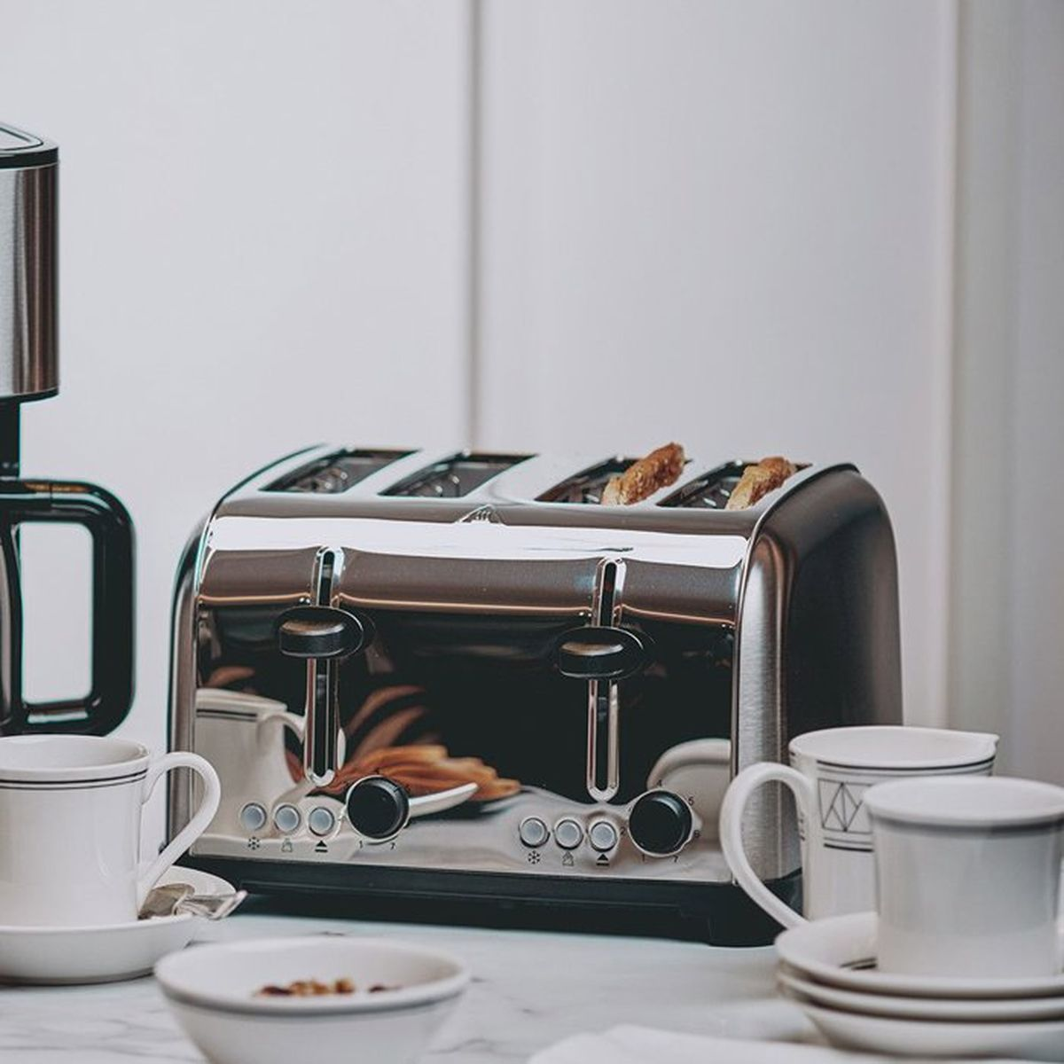Ein glänzender Toaster mit vier Toastscheiben, daneben Tassen und eine Schale.