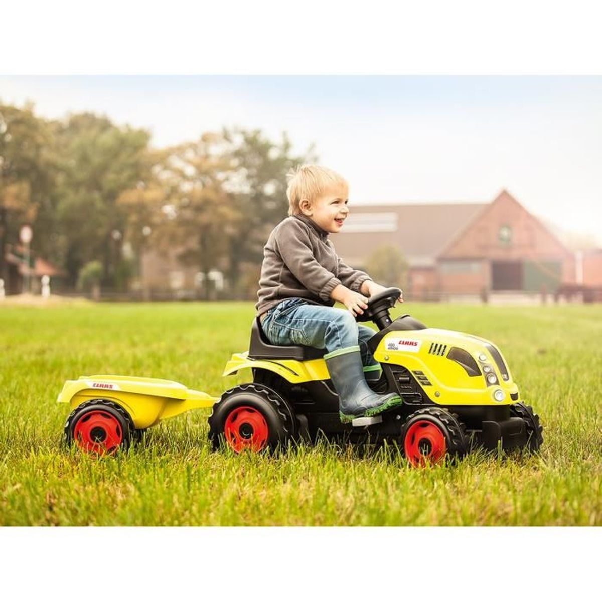 Ein Junge fährt einen gelben Spielzeugtraktor mit Anhänger auf einer grünen Wiese.