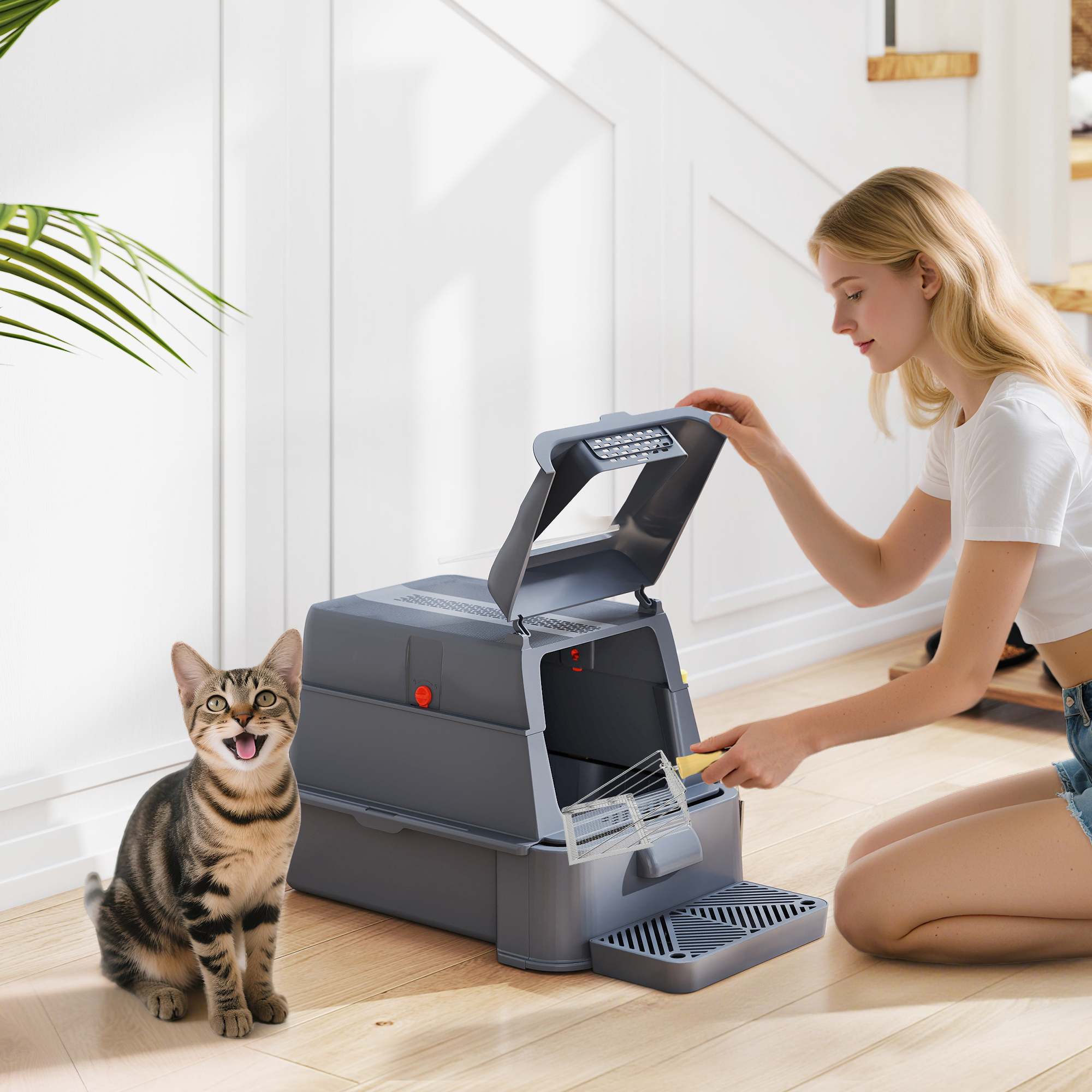 Un gato atigrado se sienta en un suelo de madera junto a una caja de arena automática que una mujer está limpiando.