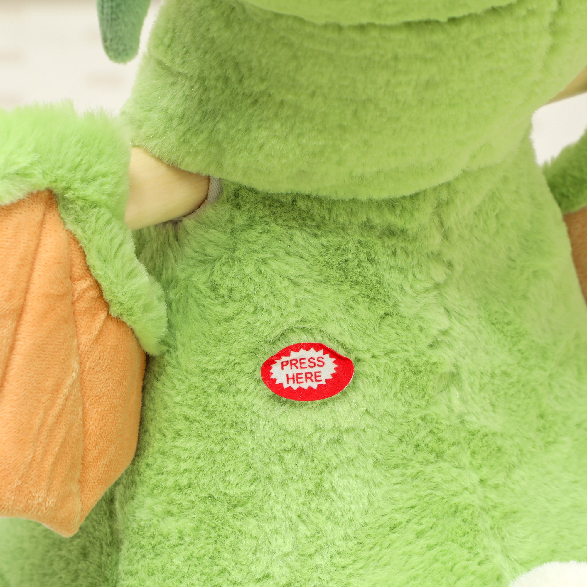 Un peluche verde con un brazo naranja y un botón rojo que dice 'Presiona aquí'.