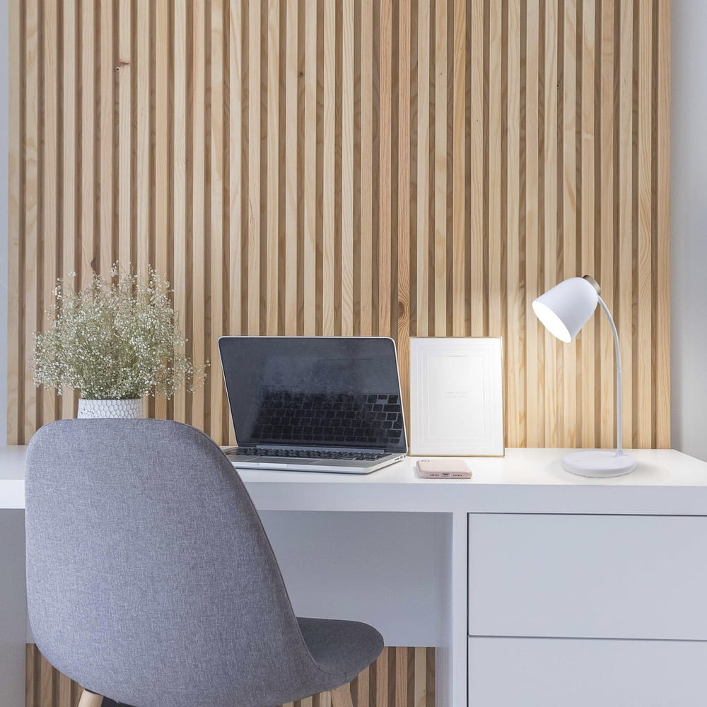 Un escritorio moderno con portátil, lámpara y planta contra pared de madera.