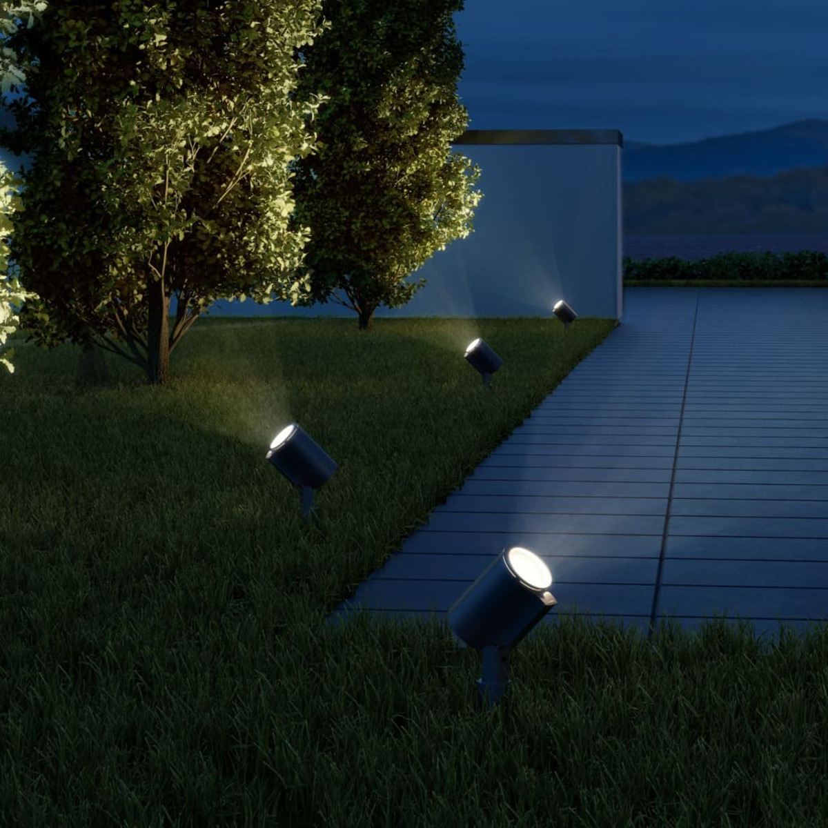 Área de césped con varios focos de suelo iluminando árboles y un camino.