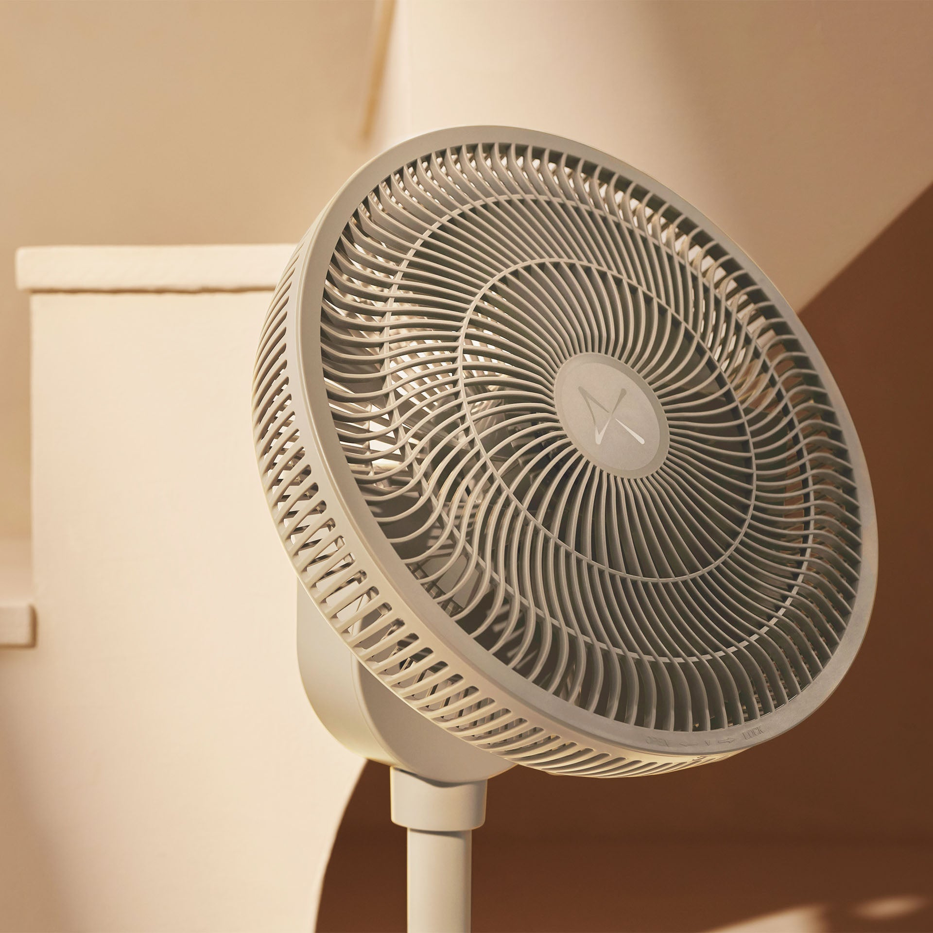 Un ventilateur sur pied gris avec une grille détaillée devant un fond beige clair.