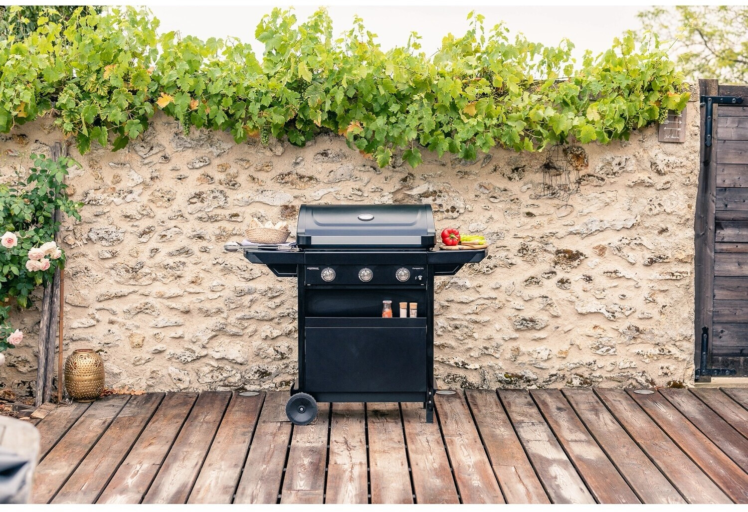 Un barbecue a gas nero su terrazza in legno davanti a muro di pietra con viti.