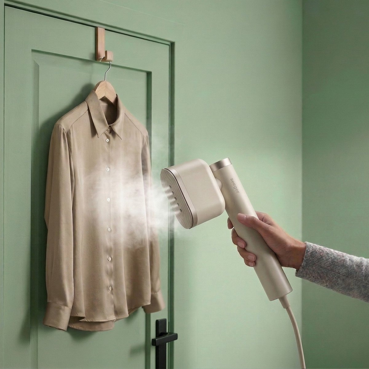 A handheld steamer smooths a beige shirt hanging on a green door.