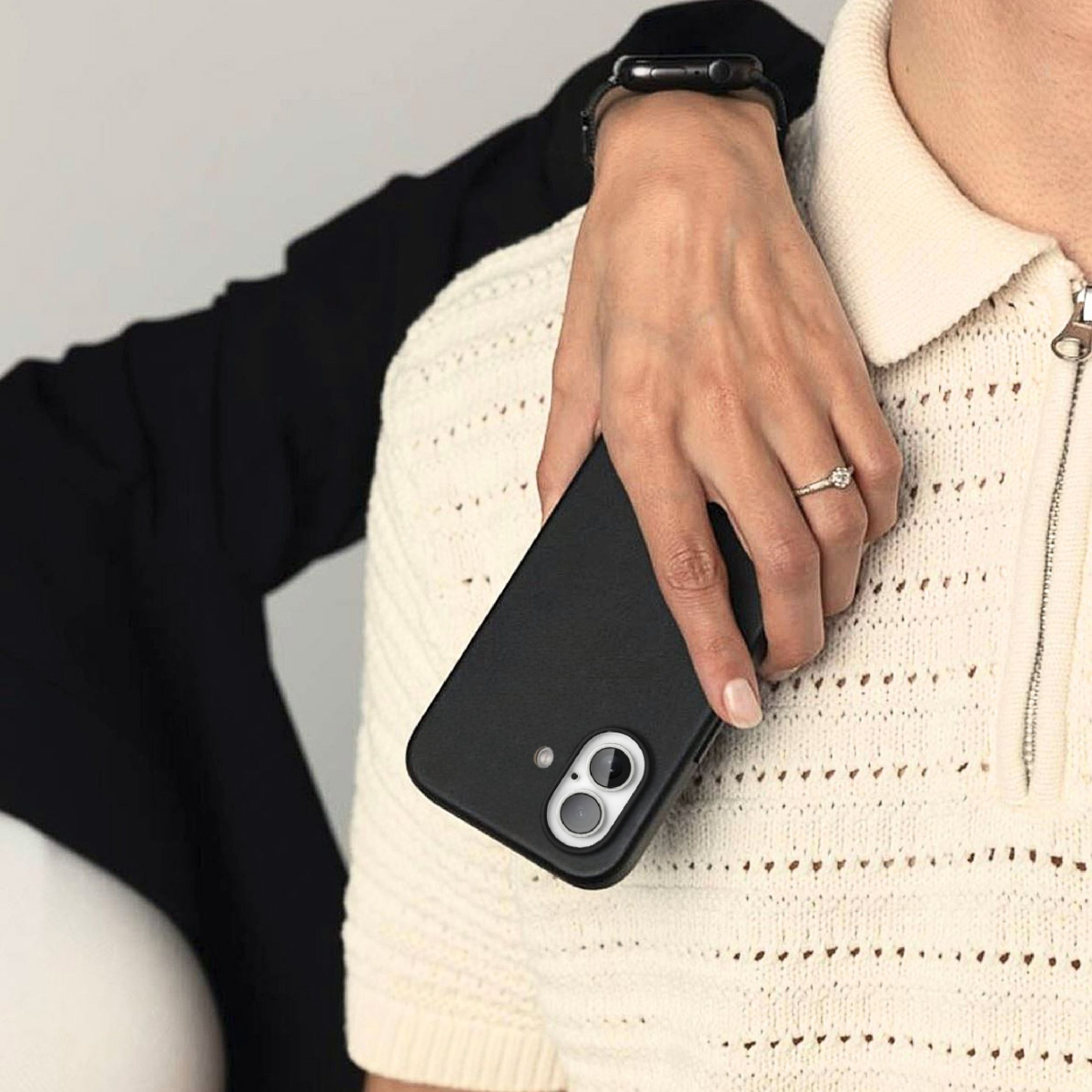 Eine Hand mit Ring hält ein schwarzes Handy vor einem cremefarbenen Pullover.