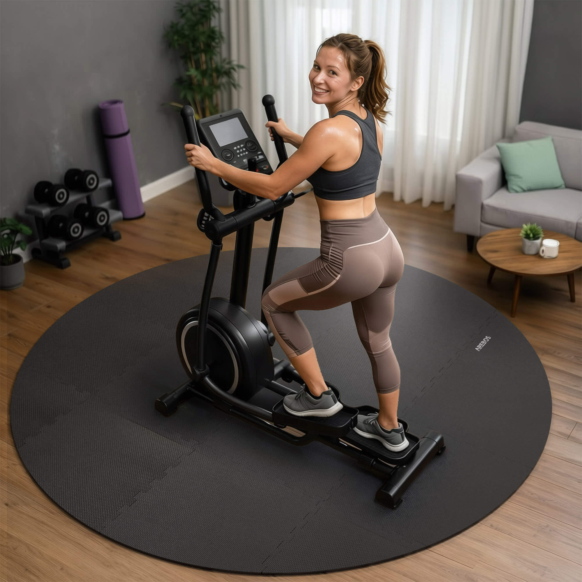 Frau trainiert auf einem Crosstrainer in einem Heim-Fitnessstudio mit Gewichten und Sofa im Hintergrund.