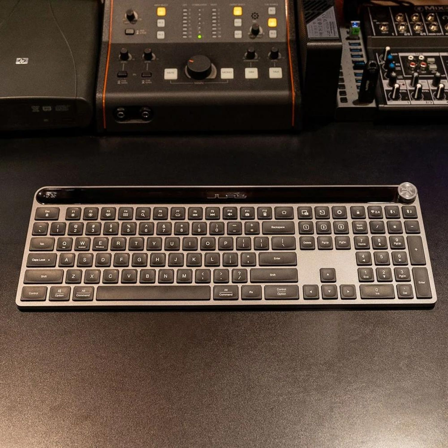 Una tastiera argentata si trova davanti a console di missaggio su una scrivania scura.