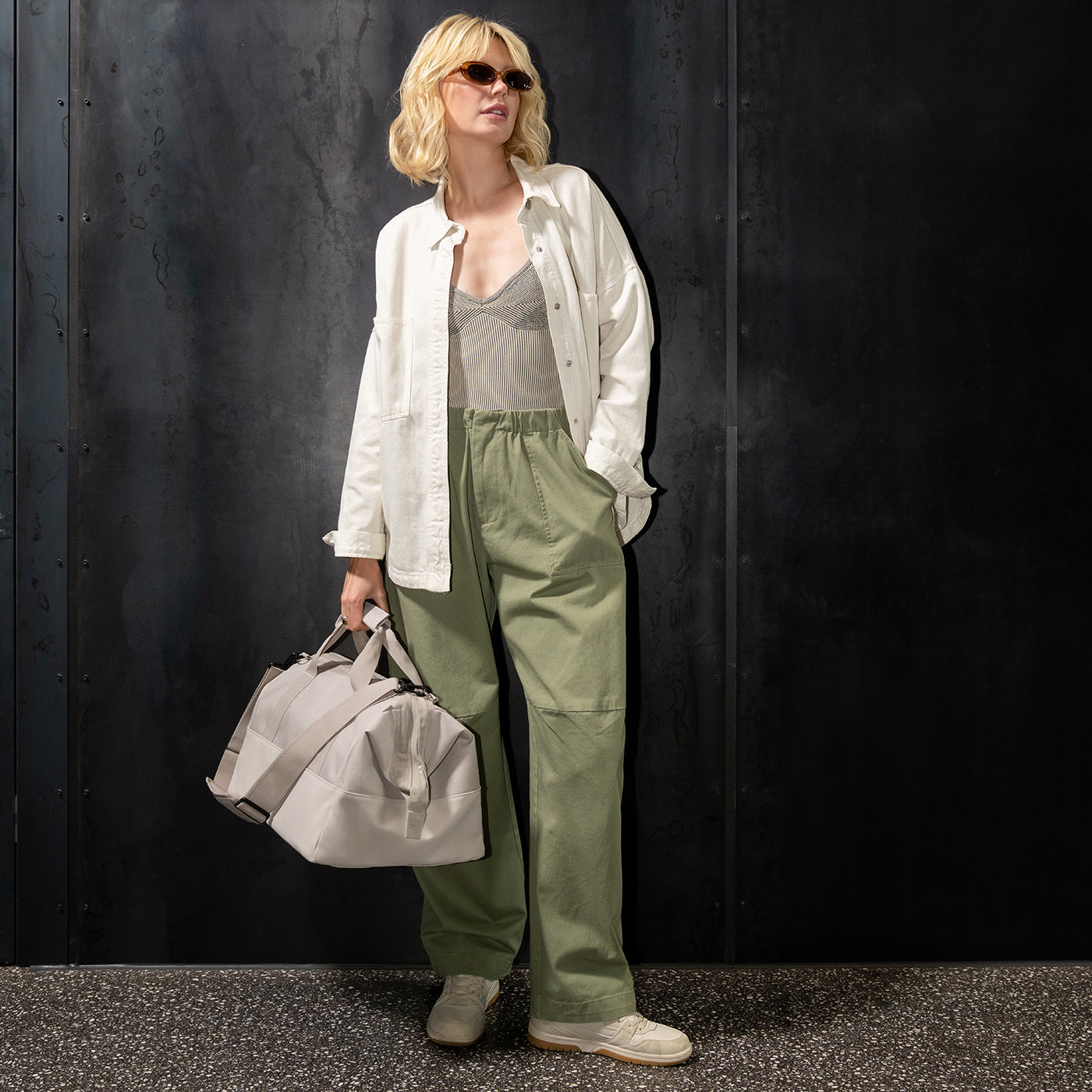 Eine Frau mit blonden Haaren und Sonnenbrille trägt eine weiße Jacke und olivgrüne Hose.