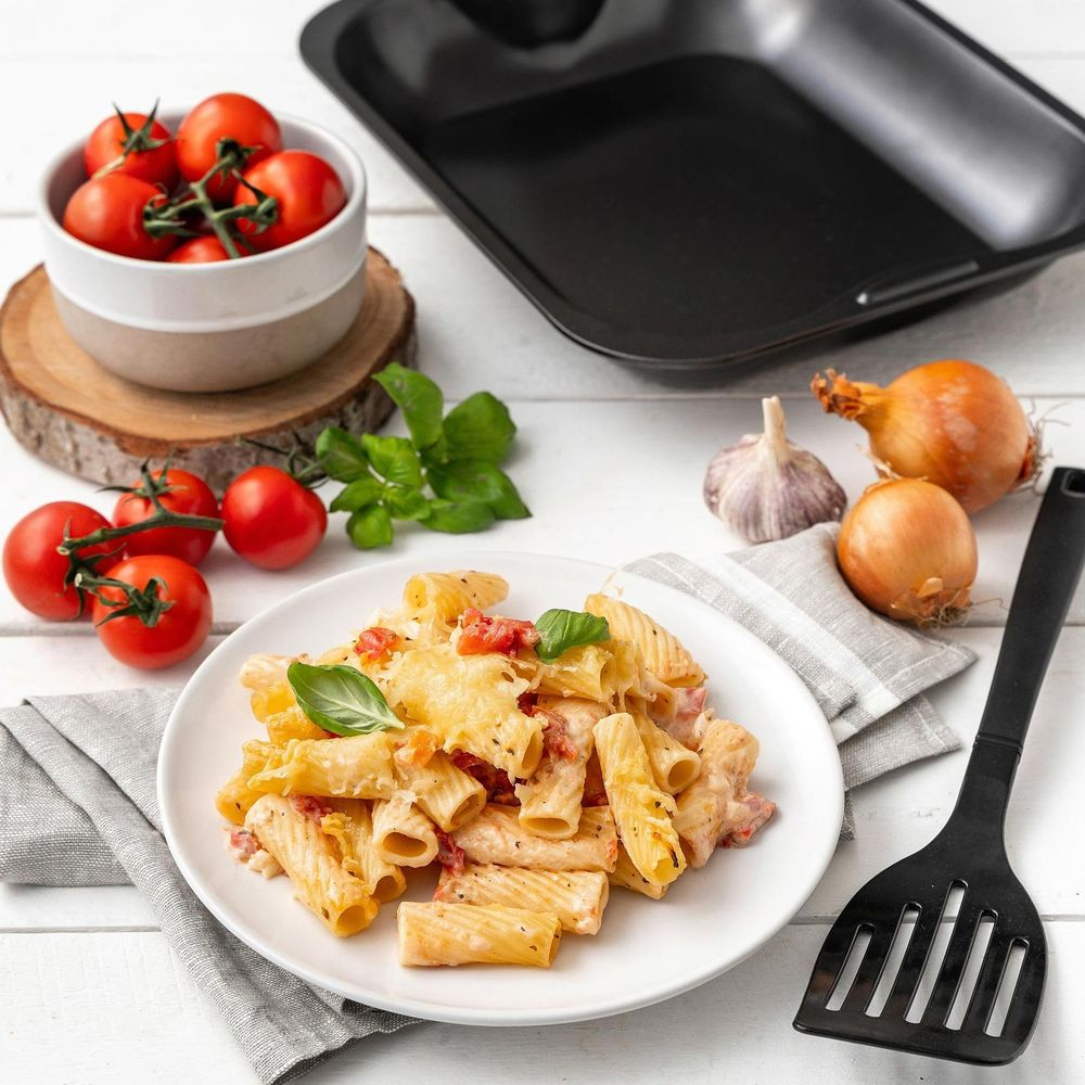 Nudelgericht mit Tomaten, Käse und Basilikum, daneben Kirschtomaten, Zwiebeln, Knoblauch und eine Bratpfanne.