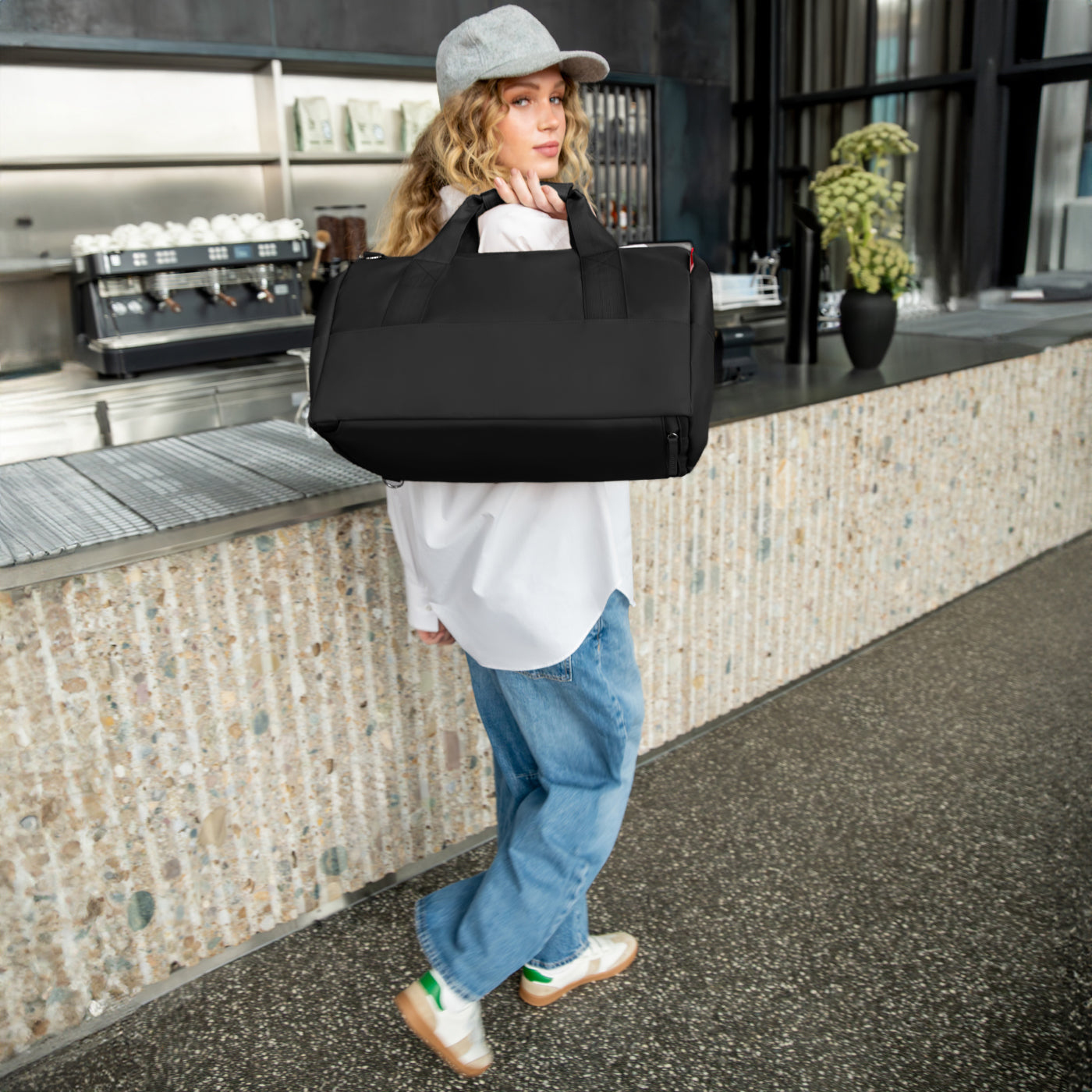 Eine Frau mit Kappe trägt eine schwarze Tasche vor einer Kaffeemaschine.