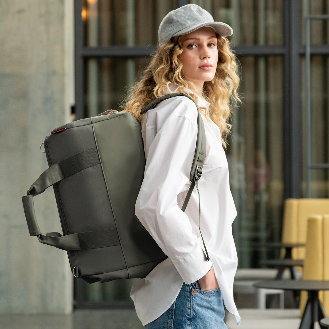 Frau mit lockigem Haar, Kappe, weißem Hemd und Jeans trägt eine grüne Tasche.