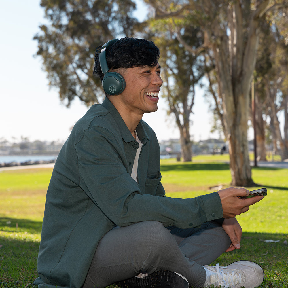 Homme avec casque est assis sur l'herbe, tenant téléphone, souriant. Arbres et eau derrière.