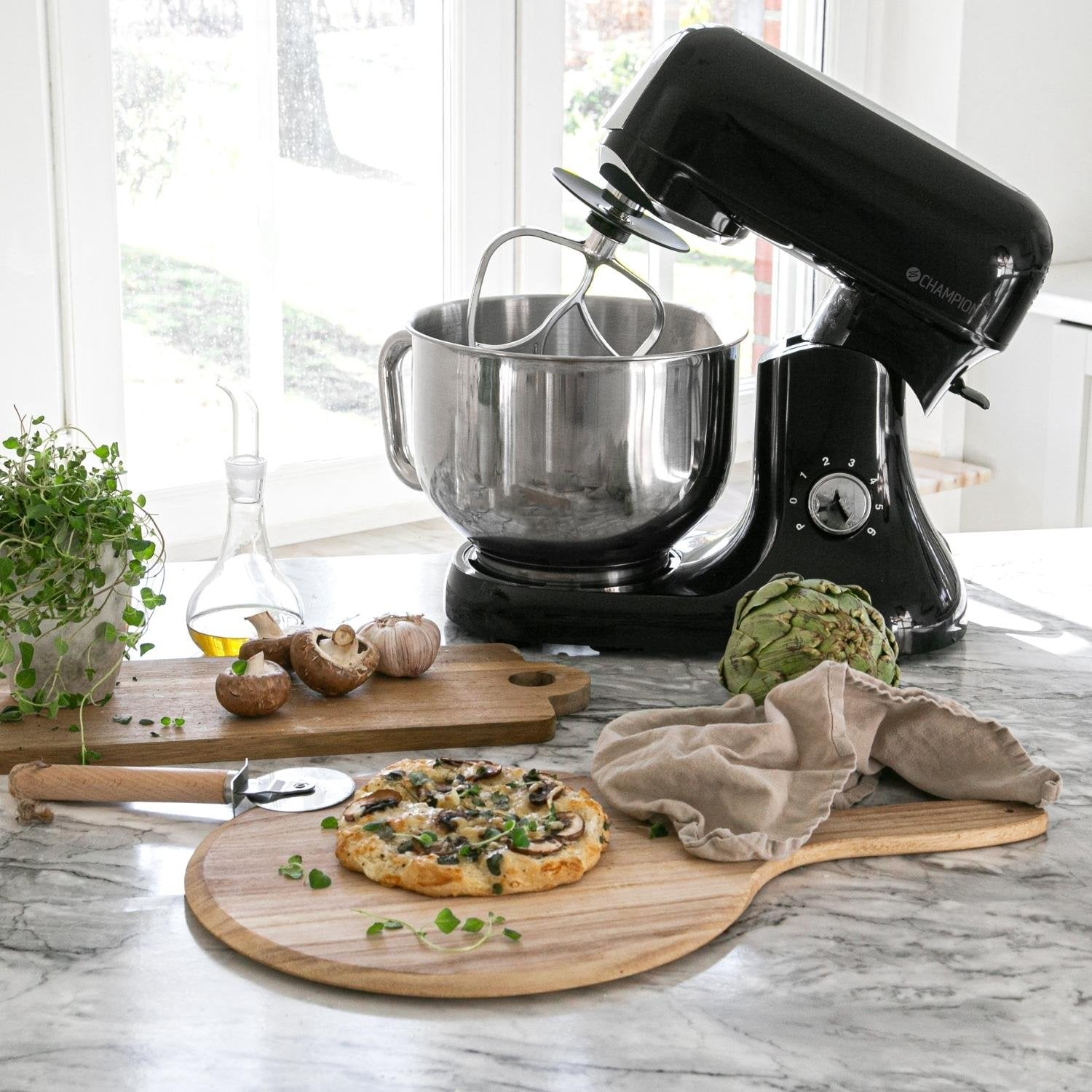 Black stand mixer with bowl, pizza, artichoke, and herbs on marble.