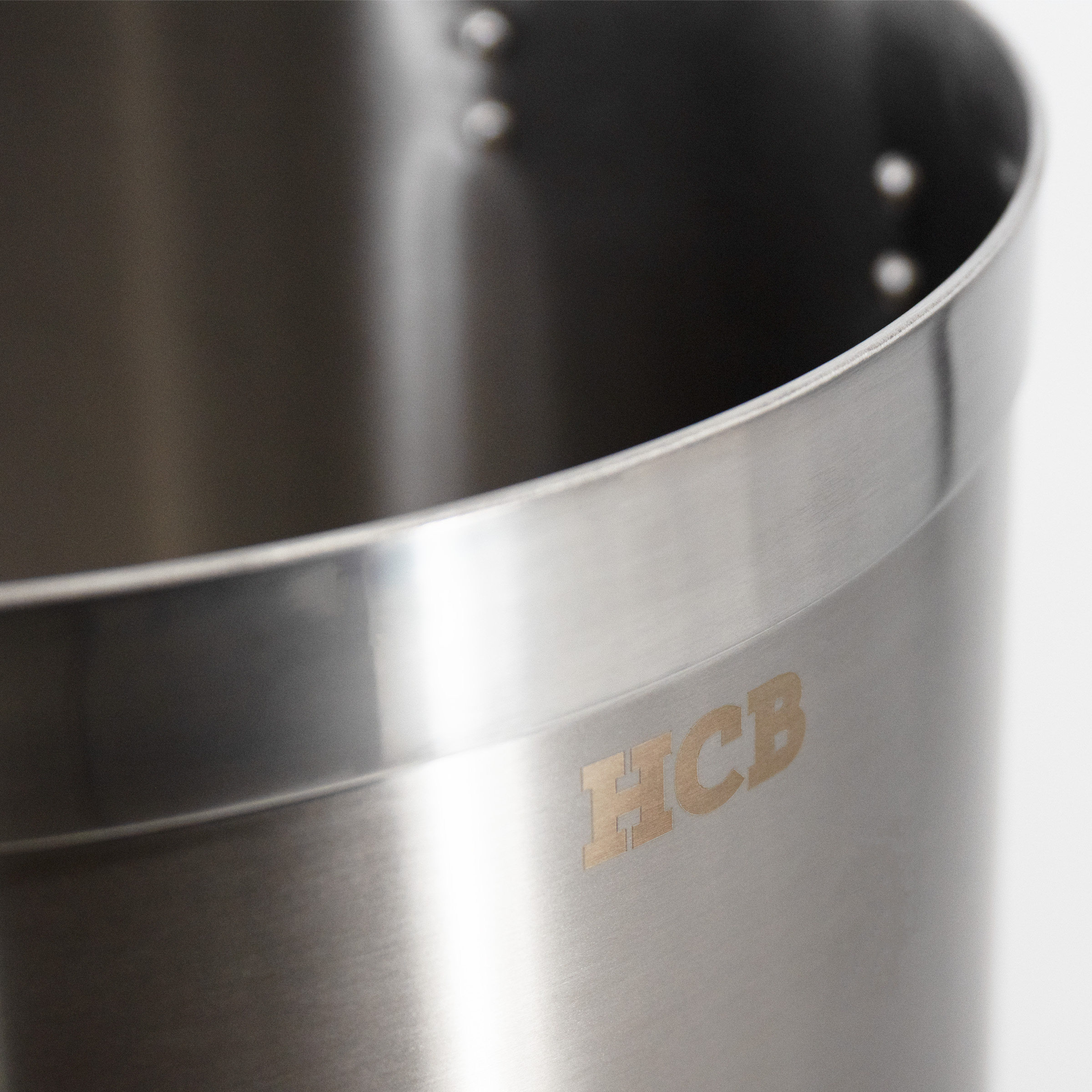Close-up van een zilveren metalen pot met het ingegraveerde HCB-logo.