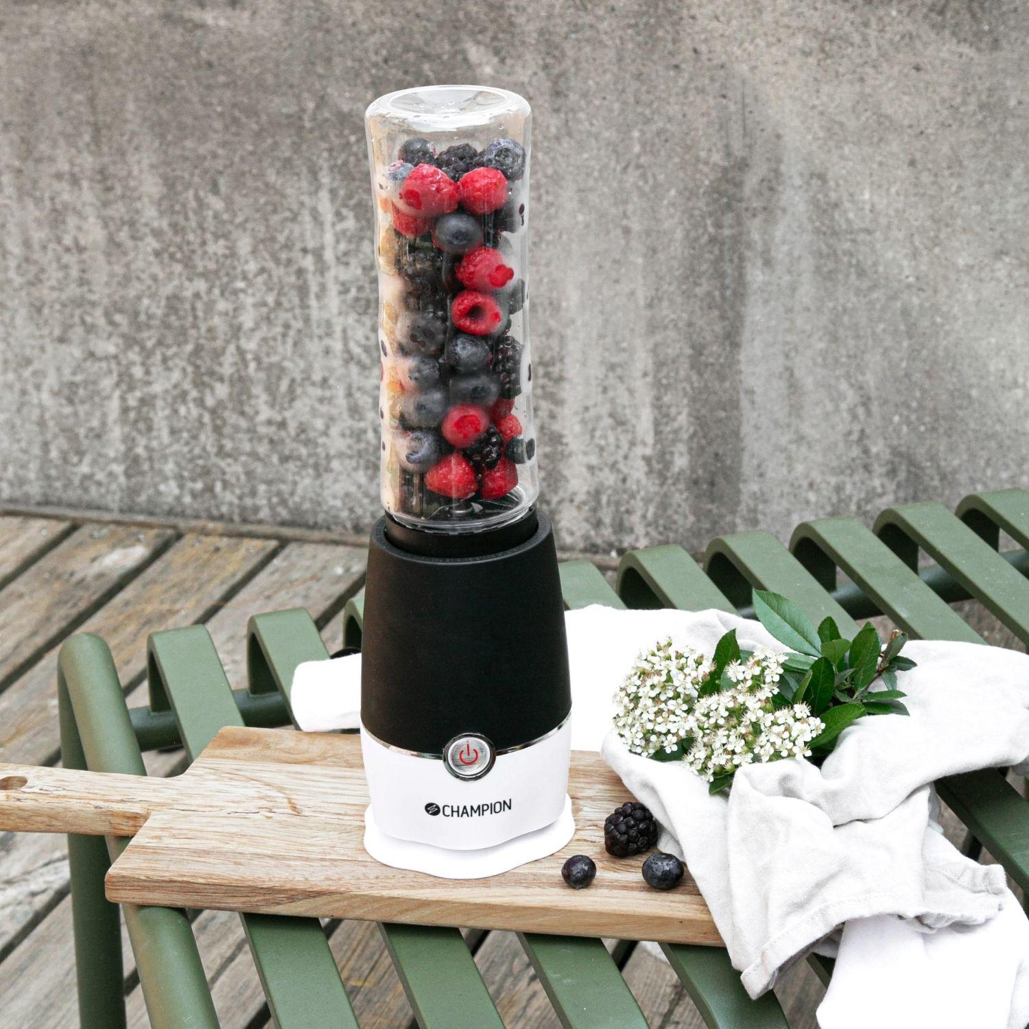 A blender filled with berries on a wooden board, next to flowers and cloths.