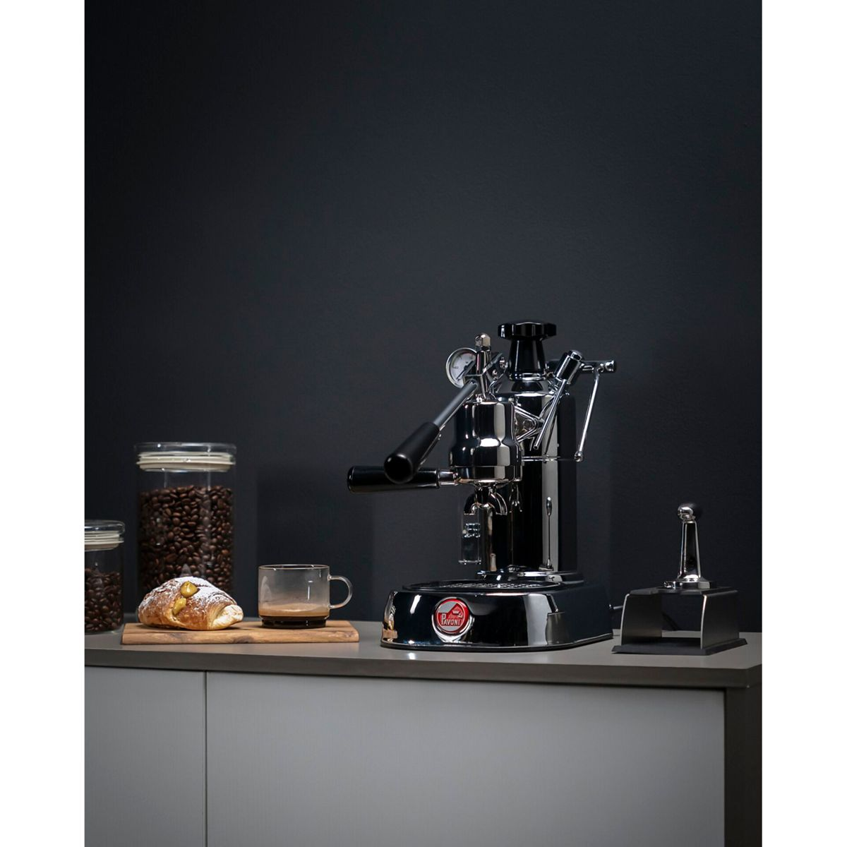 Eine Chrom-Espressomaschine steht auf einer Arbeitsplatte neben Kaffeebohnen und einem Croissant.