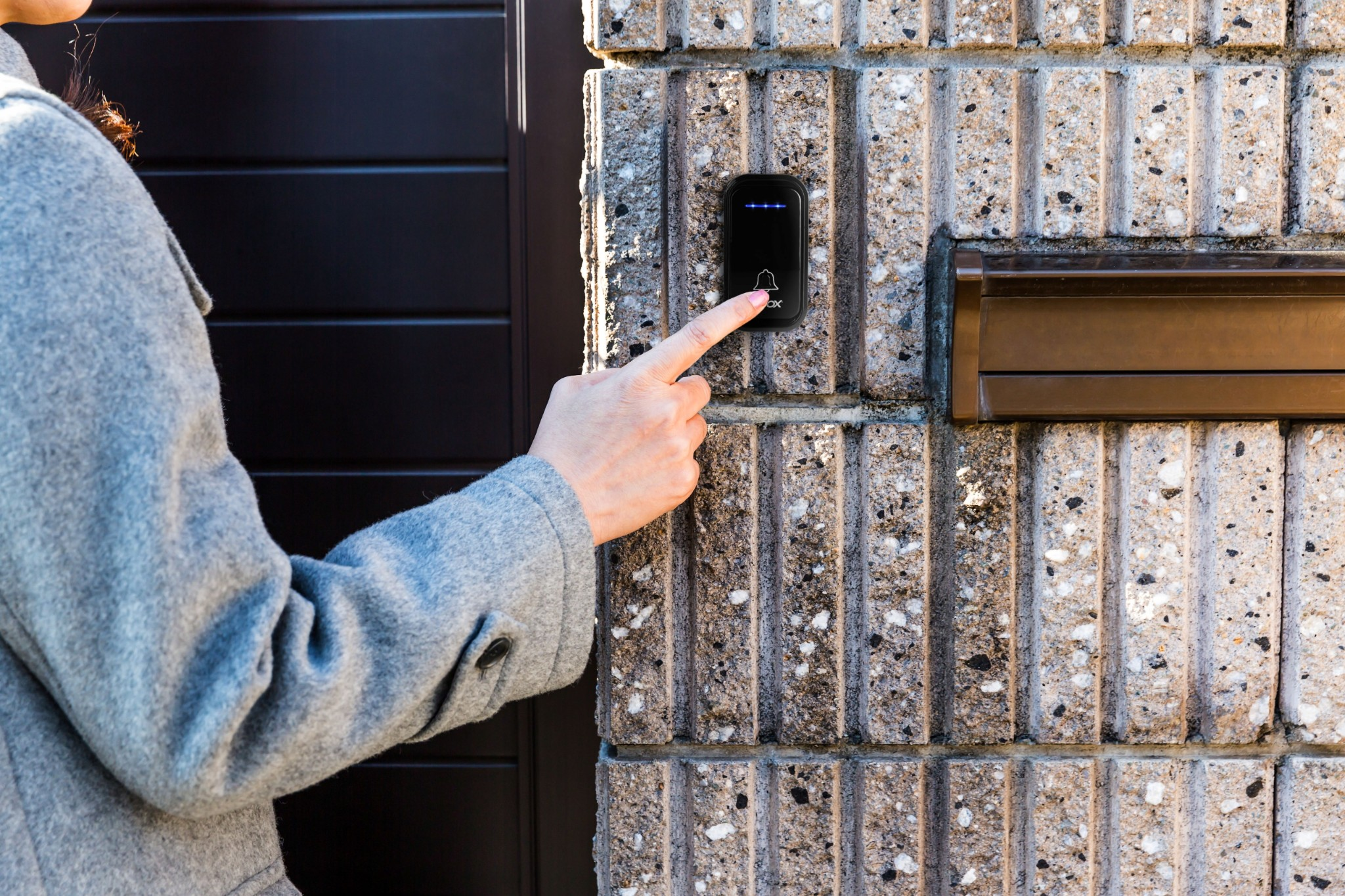 Dłoń w szarym płaszczu naciska dzwonek do drzwi na kamiennej ścianie.