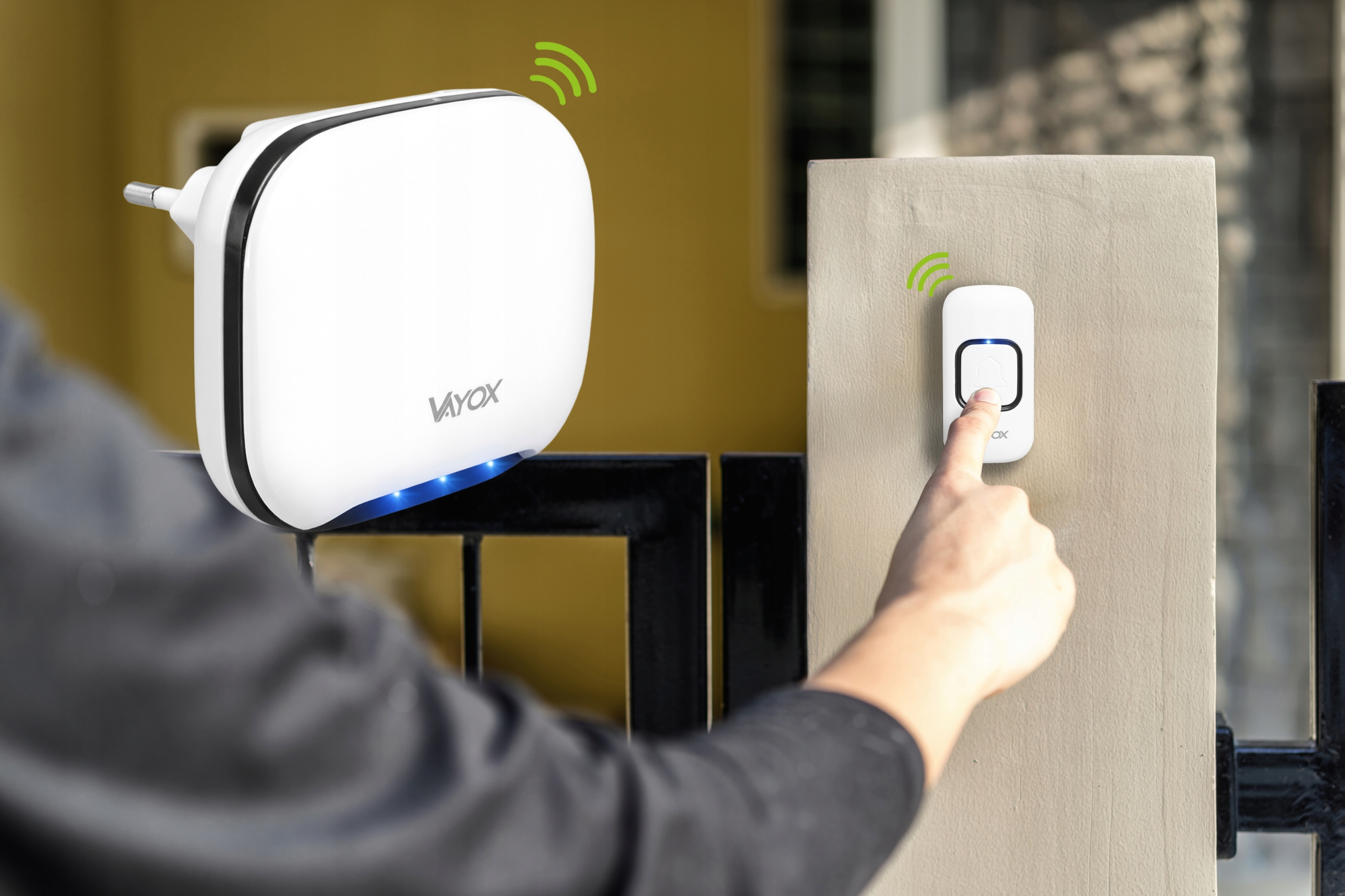 Ręka naciska biały dzwonek do drzwi, obok znajduje się biały odbiornik podłączany do gniazdka.