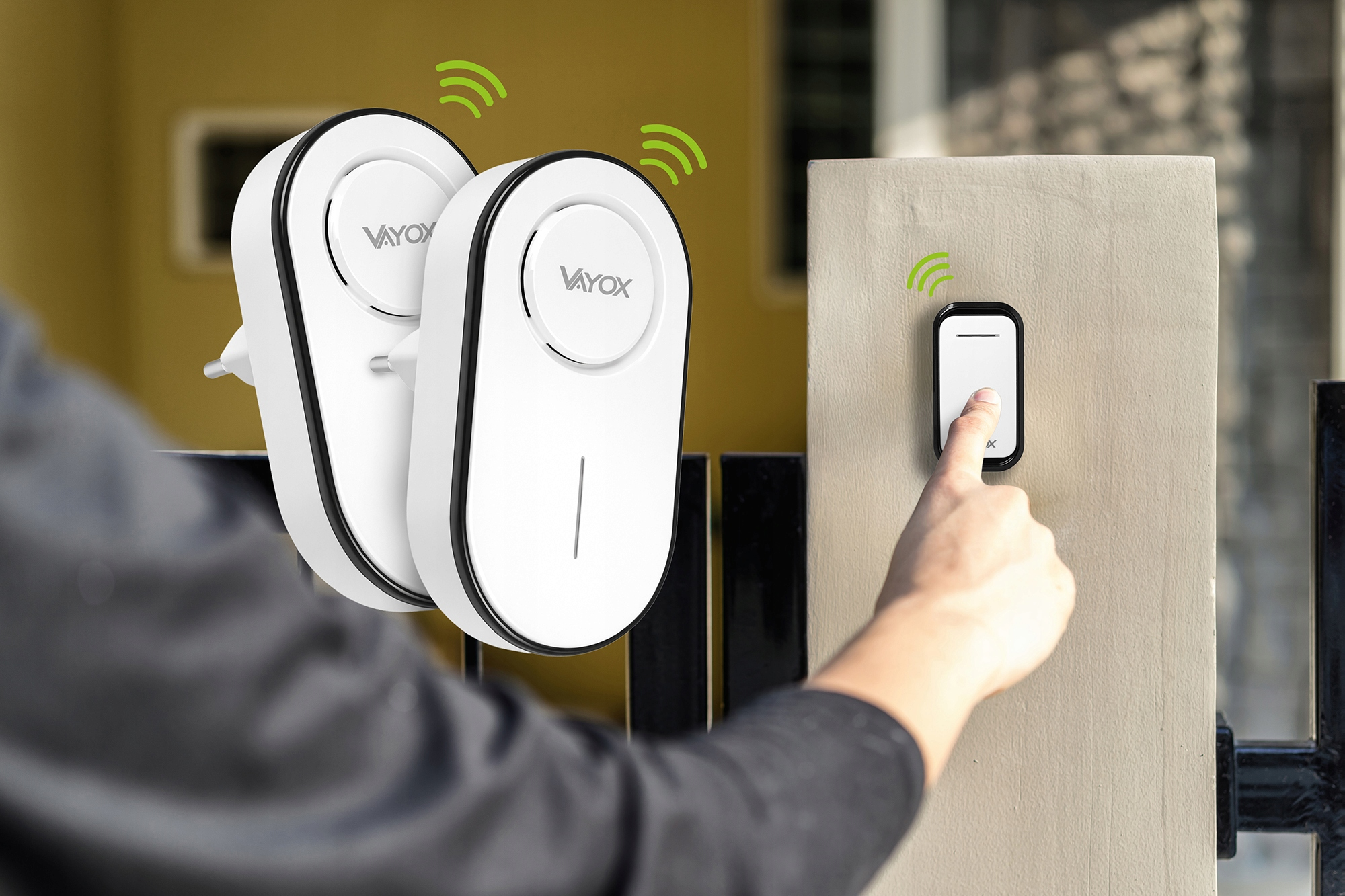 Dłoń naciska dzwonek Vayox. W pobliżu znajdują się dwa inne urządzenia.