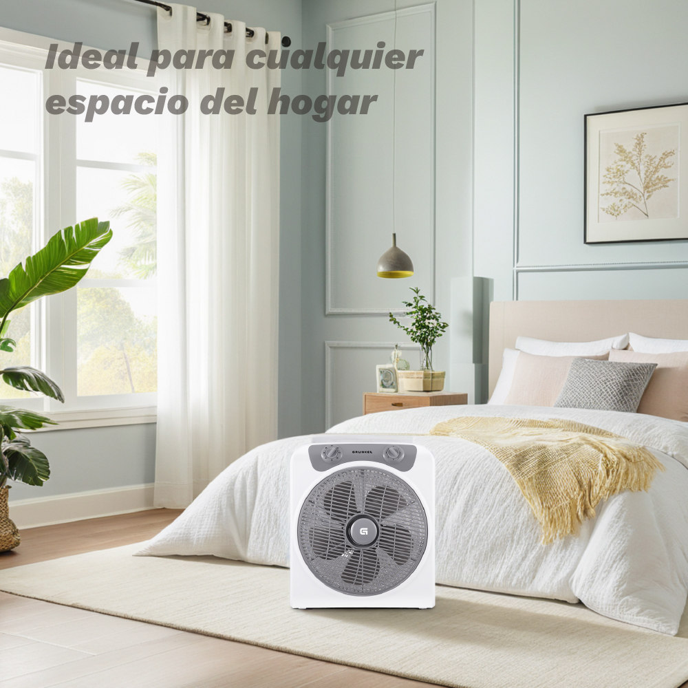 Un ventilador blanco se sienta sobre una alfombra frente a una cama en un dormitorio luminoso.